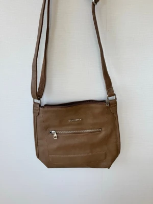 Brun axelväska från Bag Street - Snygg brun axelväska från Bag Street i slät konstläder med justerbar axelrem. Väskan har en praktisk ytterficka med dragkedja och en enkel, stilren design. Perfekt för dig som vill ha en smidig och trendig väska till vardagen.