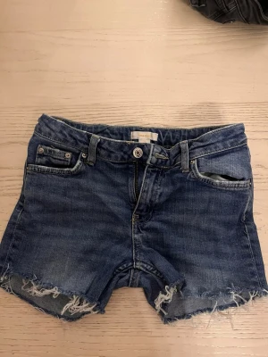 Blå jeansshorts med fransig kant - Snygga blå jeansshorts från Perfect Jeans med fransig kant nertill och klassisk femficksdesign. Shortsen har normal passform och är tillverkade i bomull. Perfekta för sommaren och enkla att matcha med olika toppar.