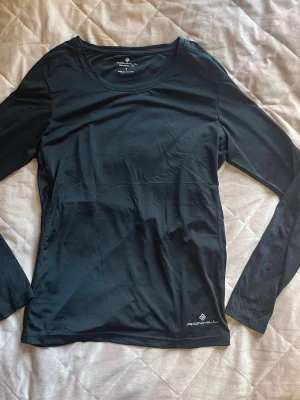 Svart långärmad träningströja Ronhill - Snygg svart långärmad träningströja från Ronhill. Tillverkad i ett lätt och stretchigt funktionsmaterial som andas bra, perfekt för löpning eller gym. Diskret logga nere vid fållen och rund halsringning för en clean look. Tror en liten söm är lös annars är den som ny!