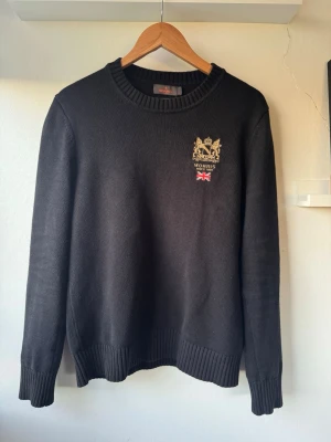 Svart stickad tröja från Morris - Svart stickad tröja från Morris med rund halsringning och ribbade muddar. På bröstet finns ett broderat emblem i guld och rött med Morris-logga och brittisk flagga. Perfekt för dig som gillar klassisk och stilren design. Den är sparsamt använd och finns där med inge defekter!! Den är i storlek S men är lite större så passar även en M