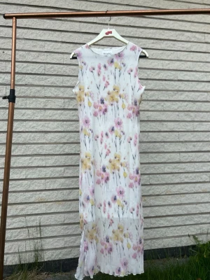 Blommig långklänning från Noella - Långklänning från Noella i vitt med blommigt mönster i pastelliga toner av rosa, gul och lila. Klänningen är ärmlös och har en rak, enkel passform. Perfekt för varma dagar och somriga tillfällen. Helt ny med prislapp, nypris: 999kr. Storlek xs.