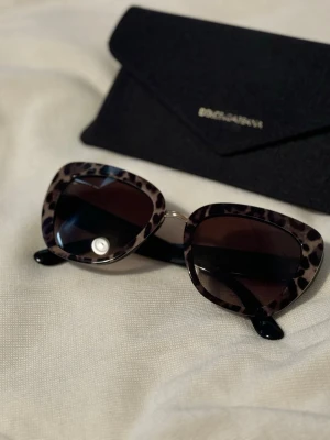 Leopardmönstrade solglasögon Dolce & Gabbana - Snygga solglasögon från Dolce & Gabbana med leopardmönstrad båge och mörka glas. Modellen har en trendig, kantig form och svarta skalmar. Perfekt accessoar för dig som vill sticka ut lite extra. Äkta solglasögon från detta varumärke 