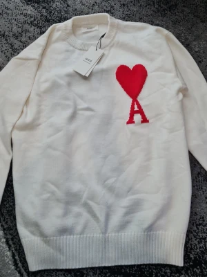 Vit stickad tröja från Ami Paris - Vit stickad tröja från Ami Paris med rund halsringning och ribbade muddar. På bröstet finns ett rött hjärta och bokstaven A, som är typiskt för märket. Perfekt för dig som gillar stilrena och trendiga plagg.