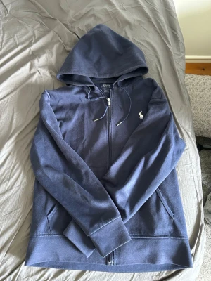 Blå hoodie från Polo Ralph Lauren - Snygg blå hoodie från Polo Ralph Lauren med dragkedja framtill och klassisk vit logga på bröstet. Hoodien har huva med dragsko och två fickor framtill. Perfekt för en avslappnad och stilren look.