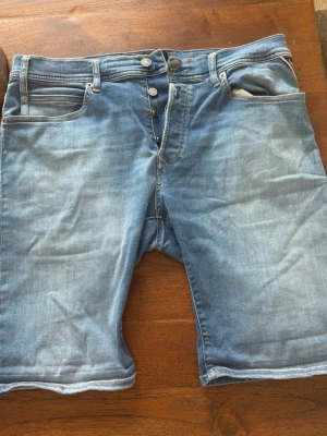 Blå jeansshorts med knappar - Snygga blå jeansshorts med klassisk femficksdesign och knäppning med flera knappar framtill. Shortsens denim har en lätt tvättad look och raka ben som slutar strax ovanför knäna. Perfekta för varma dagar och enkel att matcha med t-shirt eller hoodie.
