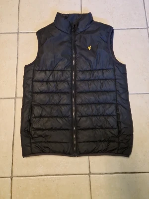 Svart dunväst från Lyle & Scott - Svart dunväst från Lyle & Scott med gul broderad logga på bröstet. Västen har ståkrage, dragkedja framtill och quiltad design med horisontella sömmar. Perfekt för lager-på-lager och enkel att matcha med olika outfits.