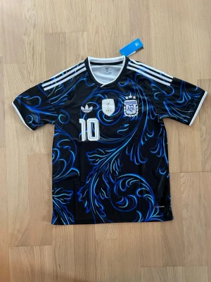 Argentina Adidas fotbollströja svart/blå - Snygg svart fotbollströja från Adidas med blått och vitt mönster, nummer 10 på bröstet och officiella Argentina AFA och FIFA 2022-emblem. Tröjan har klassiska vita Adidas-ränder på axlarna och rund hals. Perfekt för dig som älskar fotboll och vill sticka ut på planen.