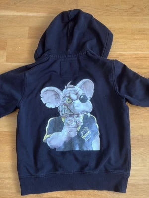 Svart AIK hoodie  - Svart hoodie med dragkedja och huva från AIK. På ryggen finns en stor tryckt bild av en tecknad råtta i AIK-tröja. AIK-logga även på huvan. Hoodien har kängurufickor och dragsko i huvan. Perfekt för dig som vill sticka ut med en unik supporterstil. 