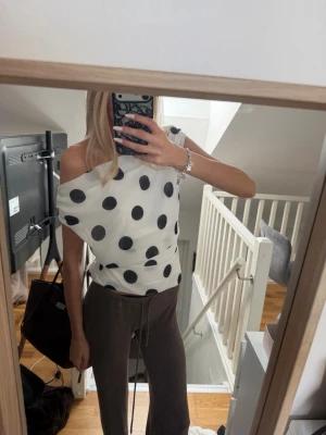 Vit prickig blus med knyt i midjan - Snygg vit blus med stora svarta prickar och trendig offshoulder-look. Toppen har knytband i midjan som ger en fin siluett och är ärmlös. Perfekt för dig som gillar en lekfull och stilren stil. xs/s