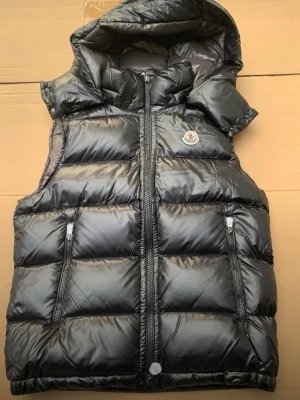 Svart dunväst från Moncler med huva - Snygg svart dunväst från Moncler med glansig finish, två dragkedjefickor och avtagbar huva. Klassisk Moncler-logga på bröstet. Perfekt för lager-på-lager och streetstyle. Fylld med dun för extra värme och komfort. 