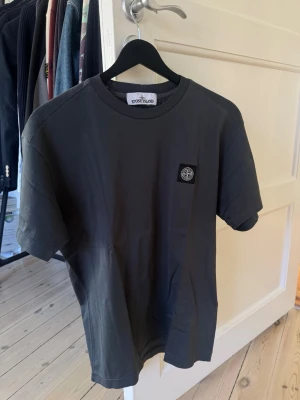 T-shirt från Stone Island - Snygg mörkgrå t-shirt från Stone Island med klassisk logga på bröstet. T-shirten har rund halsringning och korta ärmar. Perfekt för en stilren och avslappnad look.