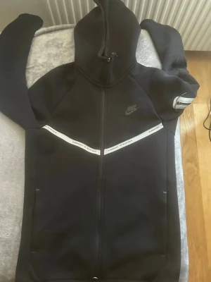 Svarta Nike mjukisbyxor med logga - Svarta mjukisbyxor från Nike med dragsko i midjan och resår vid bensluten. Byxorna har en vit rand med Nike-text längs sidorna och en diskret Nike-logga på låret. Perfekta för en avslappnad och sportig stil.
