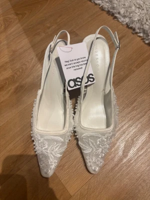 Vita spetsiga klackar med broderi - Eleganta vita pumps från ASOS DESIGN med spetsig tå, smal klack och justerbar rem. Skorna är dekorerade med vackert blommigt broderi och pärldetaljer på ovandelen. Perfekta till bal eller student. De är helt oanvända och slutsålda på hemsidan. Klackhöjd 6 cm.