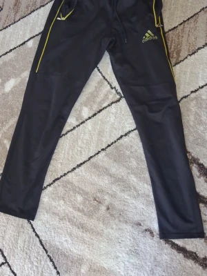 Svarta Adidas mjukisbyxor med gula detaljer - Svarta mjukisbyxor från Adidas med gula dragkedjor och logga på vänster ben. Byxorna har smal passform, resår i midjan och dragsko för bästa komfort. Perfekta för chill eller träning, tillverkade i mjukt och följsamt material.