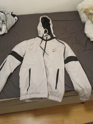 Nike grå och svart vindjacka med huva - Snygg grå vindjacka från Nike med svarta detaljer och dragkedjor. Jackan har huva med dragsko, två fickor med dragkedja och svart meshfoder på insidan. Perfekt för blåsiga dagar och sportiga outfits.
