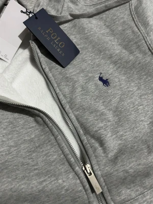 Grå hoodie med dragkedja från Polo Ralph Lauren - Snygg grå hoodie från Polo Ralph Lauren med dragkedja och blå broderad logga på bröstet. Tillverkad i mjukt material med vit fleece på insidan och praktisk huva. Perfekt för en avslappnad och stilren look. Ny med prislapp