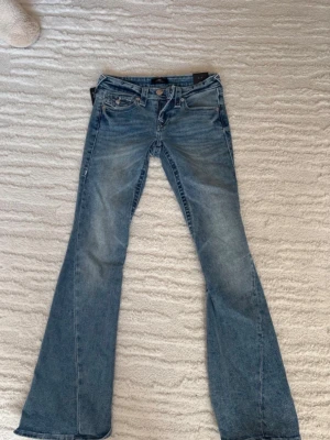Blå bootcut jeans från True Religion - Snygga blå bootcut jeans från True religion med  dekorativa bakfickor, broderi och knappar. Klassisk femficksmodell i denim med låg midja och utsvängda ben.  Storlek 25. Aldrig använda prislapp kvar 