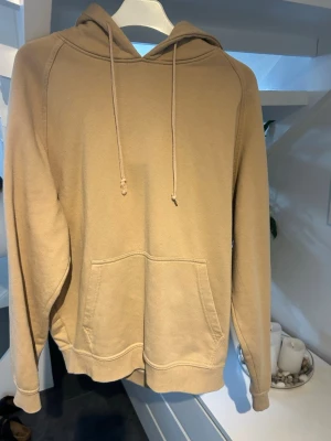 Beige hoodie med ficka framtill - Snygg beige hoodie med huva och dragsnören. Hoodien har en stor känguruficka framtill och ribbade muddar vid ärmslut och nederkant. Tillverkad i mjukt sweatshirtmaterial, perfekt för en avslappnad stil.