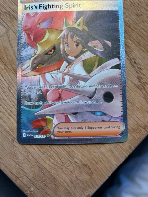 Pokémonkort Iris's Fighting Spirit - Säljer ett holografiskt Pokémonkort med motivet Iris's Fighting Spirit. Kortet har färgstarka illustrationer med blå, röd och gul ton samt detaljerad design. Perfekt för samlare eller Pokémonspelare som vill utöka sin kortlek.