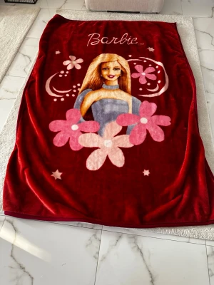 Röd Barbie filt med blommor - Mysig röd filt med stor Barbie-illustration och rosa blommor. Filtens motiv har även stjärnor och dekorativa detaljer i vitt och rosa. Perfekt för att mysa i soffan eller som dekoration i sovrummet. Materialet är mjukt och lent syntet.