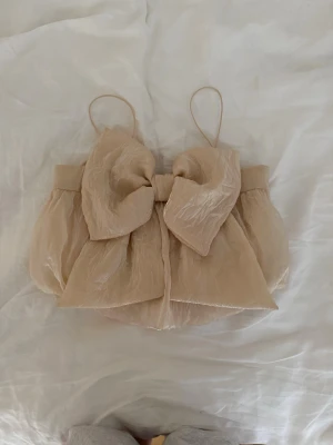 Beige linnetopp med stor rosett - Supersöt beige linnetopp med tunna axelband och en stor rosett framtill. Toppen har en luftig och lätt struktur med voluminös form, perfekt för en trendig look. Materialet känns skirt och ger en romantisk vibe.