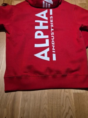Röd hoodie från Alpha Industries - Riktigt snygg hoodie i Speed Red från Alpha Industries med stor vit logga på ryggen och på huvan. Dragsko i huvan och ribbade muddar och ficka på magen. Perfekt för dig som gillar streetwear och vill sticka ut lite extra. Köpt i vintras på zalando för 950kr men har blivit hängande oanvänd endast prövad.