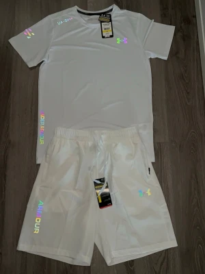 Under armour set M - Snyggt vitt träningsset från Under Armour med t-shirt och shorts. Setet har holografiska loggor och textdetaljer i regnbågsfärger. Materialet är lätt och andas, perfekt för sport och träning. Elastisk midja på shortsen för skön passform.