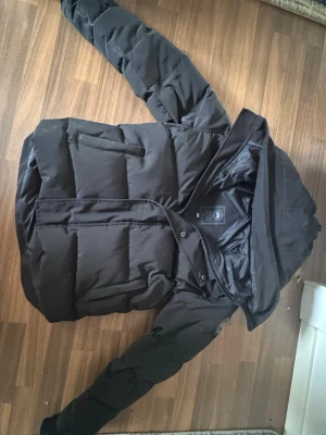 Canada Goose Jacka - Säljer en svart pufferjacka från Canada Goose med klassisk patch på ärmen. Jackan har huva, dragkedja och tryckknappar framtill samt breda, vadderade paneler för extra värme. Perfekt för kalla dagar och riktigt snygg streetstil.