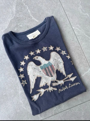 Denim&supply Ralph Lauren - Mörkblå t-shirt med broderad örn och stjärnor på bröstet samt Ralph Lauren-logga. T-shirten har rund halsringning och korta ärmar. Snygg detalj med amerikanskt inspirerat motiv i vitt, rött och blått. Perfekt för en avslappnad och cool stil.