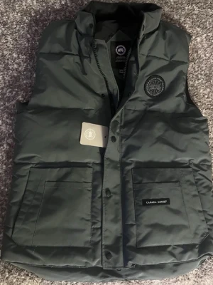 Mörkgrön dunväst från Canada Goose - Stilren mörkgrön/mörkgrå dunväst från Canada Goose med hög krage, två stora framfickor och diskret logotyp på bröstet. Västen har en klassisk, rak passform och stängs med både dragkedja och tryckknappar. Perfekt för lager-på-lager under kyliga dagar.
