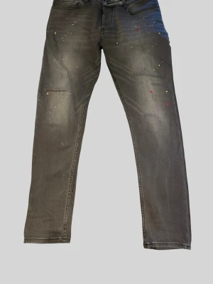 Grå jeans ifrån Dsquared2 med färgstänk  - Snygga grå jeans med raka ben och coola färgstänk i rött och vitt. Något mer slimfit än regular utan att kännas för tighta. Jeansen har klassisk femficksdesign och subtila slitningar framtill för en trendig look. Perfekta för dig som vill sticka ut lite extra i vardagen, design typiskt för märket Dsquared2. Inga tecken på användning framstår som helt nya. Säljes långt under halva nypriset. 