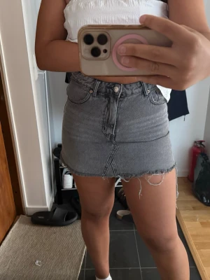 Grå jeanskjol från Gina Tricot - Trendig grå jeanskjol från Gina Tricot med rå kant och fransar nertill. Kjolen har klassisk femficksdesign, knapp och dragkedja framtill samt en kort passform. Perfekt för dig som gillar en avslappnad och cool stil.