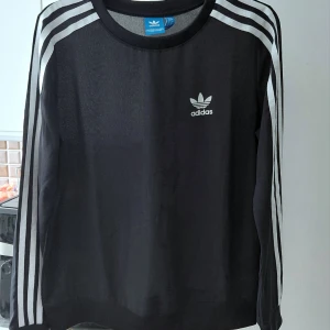 Adidas Sweatshirt,  34/36 - Svart långärmad tröja från Adidas med den klassiska Trefoil-logotypen på bröstet. Tröjan har tre vita ränder längs ärmarna, från axel till mudd. Muddarna är ribbade för en bekväm passform. Halsringningen är rund. Från armhålan till armhålan 50cm/utan elastan!/Knappt använd.