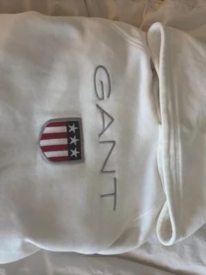 Vit hoodie från GANT med logga - Snygg vit hoodie från GANT med stor broderad logga och amerikanskinspirerat sköldmärke på bröstet. Är använd ett fåtal gånger! Skriv om du ha några frågor eller vill ha fler bilder!💗