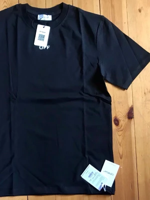 Svart Off-White t-shirt med tryck - Svart t-shirt från Off-White med liten vit 'OFF'-logga tryckt på bröstet. Klassisk rund hals och korta ärmar. Tillverkad i mjuk bomull för en skön känsla. Perfekt för en clean och stilren look.