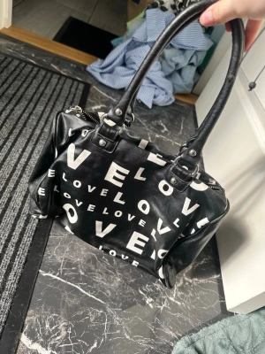 Svart handväska med LOVE-tryck - Cool svart handväska med stora vita bokstäver som stavar 'LOVE' över hela väskan. Väskan har två rejäla handtag och är tillverkad i ett glansigt konstlädermaterial. Perfekt för dig som vill sticka ut med en statement-väska.