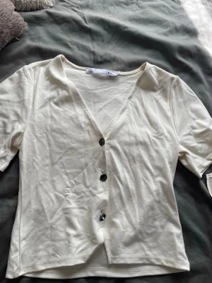 Vit kortärmad topp med knappar - Snygg vit topp med v-ringning och tre dekorativa knappar framtill. Kortärmad och croppad modell i mjukt bomullsmaterial. 