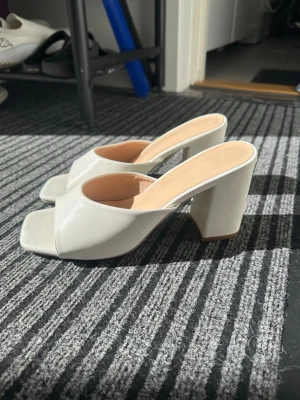 Vita chunky slip-in pumps - Stilrena vita pumps med öppen tå och bred klack. Skorna har slip-in-modell med fyrkantig tå och är tillverkade i ett glansigt material som ger en modern look. Perfekta för dig som gillar trendiga och bekväma klackar.