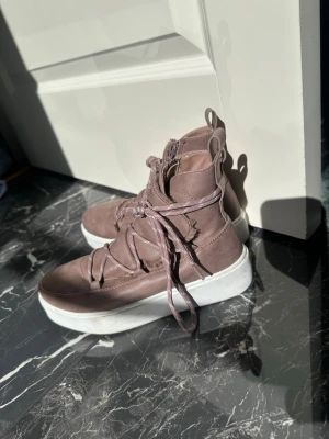 Ljuslila höga sneakers med platåsula - Snygga höga sneakers i ljuslila mockaliknande material med bred vit platåsula. Skorna har snörning framtill, dragkedja på sidan och praktiska dragflikar både fram och bak. Perfekta för dig som vill sticka ut med en unik stil.