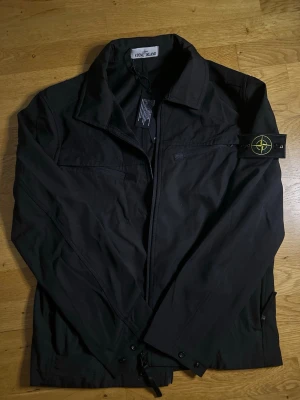 Svart overshirt från Stone Island - Säljer en svart overshirt från Stone Island med klassisk krage och dragkedja framtill. Jackan har den ikoniska logotypen på vänster ärm och är tillverkad i ett slitstarkt material. Perfekt för lager-på-lager och streetwear-stil.