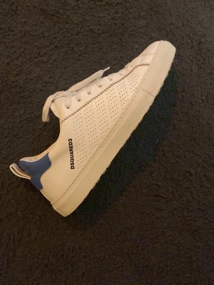 Vita DSQUARED2 sneakers med blå detalj - Snygga vita sneakers från DSQUARED2 med perforerade sidor och blå detalj vid hälen. Klassisk låg modell med vita skosnören och robust sula. Perfekta för dig som gillar stilrena och trendiga skor.