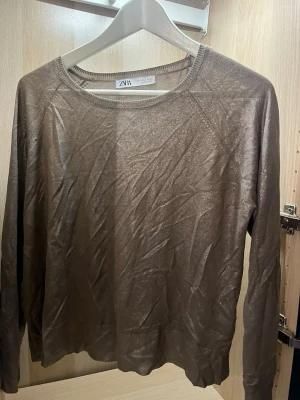 Brons/guld färgad tunn tröja från ZARA - Snygg tunn långärmad tröja från ZARA i skimrande brons/guld färg  Tröjan har rund halsringning och är tillverkad i ett lätt, glansigt material som ger en trendig look. Perfekt att styla lager på lager eller som den är. Använd mycket få gånger!