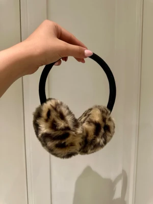 Leopardmönstrade öronmuffar - Snygga öronmuffar med leopardmönster i beige, brunt och svart. De är gjorda i mjuk fuskpäls och har ett svart band som sitter bekvämt över huvudet. Går att lyssna på musik med de om man har rätt sladd.