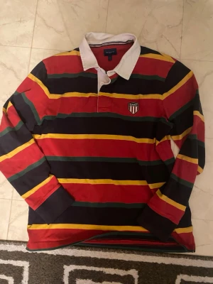 Randig rugbytröja från GANT - Snygg långärmad rugbytröja från GANT med breda ränder i rött, gult, grönt och marinblått. Kolla sista bilden!