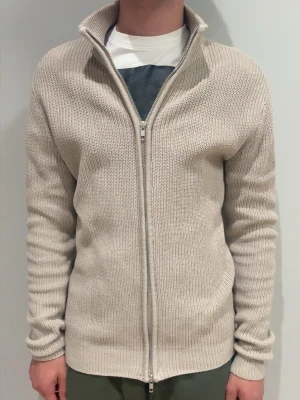 Beige stickad ziptröja med hög krage - Snygg beige stickad tröja med dragkedja och hög krage. Tröjan har ribbstickad struktur och långa ärmar, perfekt för lager-på-lager. Enkel och stilren design som passar till det mesta. Hör av er gällande frågor!!