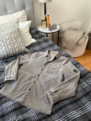 Cp Company grå overshirt  - Säljer en snygg grå overshirt i manchestertyg med klassisk krage, bröstficka och stora knappar. Overshirten har en avslappnad passform och detaljer på ärmarna, perfekt för lager på lager. Passar dig som gillar stilrena och tidlösa plagg.