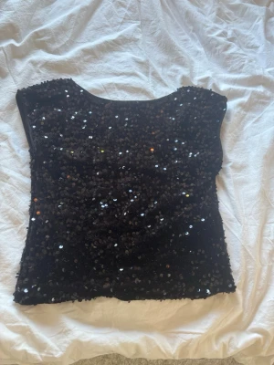 Svart paljettopp med rund hals - Snygg svart topp täckt med glittrande paljetter. Modellen är ärmlös med rund halsringning och croppad passform. Perfekt för dig som vill sticka ut och glänsa lite extra. Materialet känns mjukt och stretchigt, troligen polyester. Tröjan är inte använd🤗