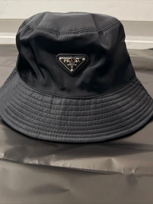 Svart bucket hat - Stilren svart bucket hat med ikonisk metallogo framtill. Hatten är tillverkad i ett glansigt syntetmaterial som ger en modern och lyxig känsla. Perfekt accessoar för att lyfta din outfit med en exklusiv touch.