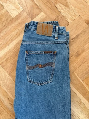 Blå raka jeans från Nudie Jeans - Klassiska blå jeans från Nudie Jeans med rak passform och hög midja. Jeansen har fem fickor, orangea sömmar och Nudie-logga på bakfickan samt läderpatch i midjan. Tillverkade i 100% bomull för en autentisk denimkänsla.