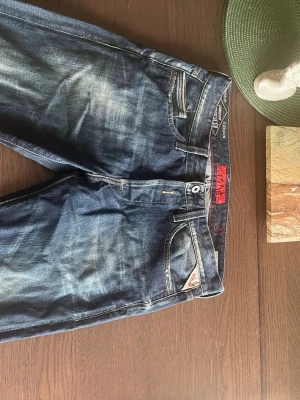 Blå Replay jeans med slitningar - Snygga blå jeans från Replay med coola slitningar och tvättade detaljer. Klassisk femficksmodell med logotyp på bakfickan och röd detalj vid linningen. Perfekta för en avslappnad och trendig stil.